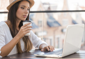 Working outdoors. Beautiful young woman in funky hat working on laptop and smiling while sitting outdoors